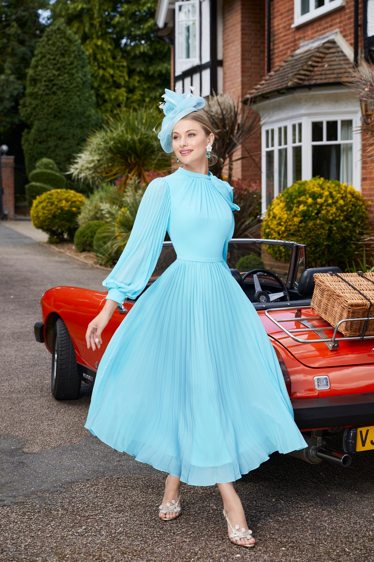 Blonde lady standing outside house next to vintage car wearing occasion a-line dress with high neck and long sleeves with sparkle heeled shoes and matching fascinator hat 